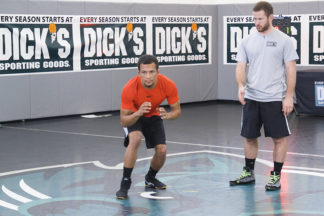 A wrestler in an orange shirt shows movement in his stance while an instructor watches on